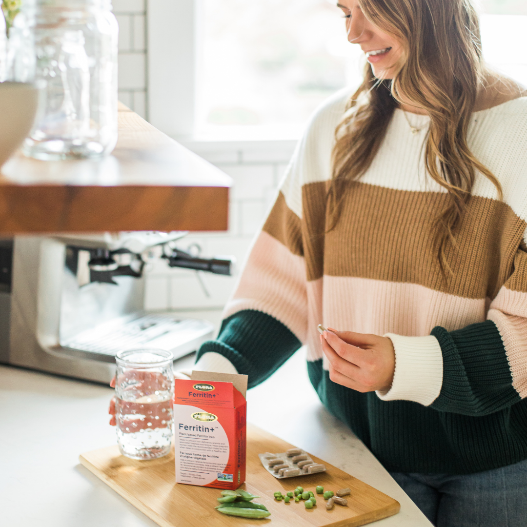Young woman holding a bottle of water with the Ferritin+ pill on her other hand and the package of the product right in front of her in a table next to some peas